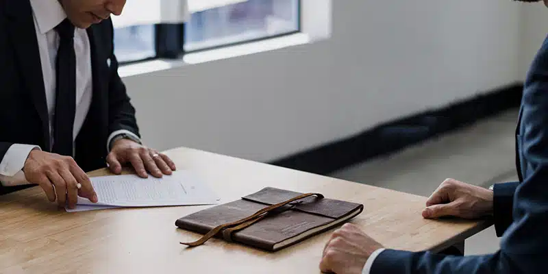 Two people in suits sit across from each other at a table; one is reviewing a financial analysis document while a closed notebook with a strap lies on the table between them.